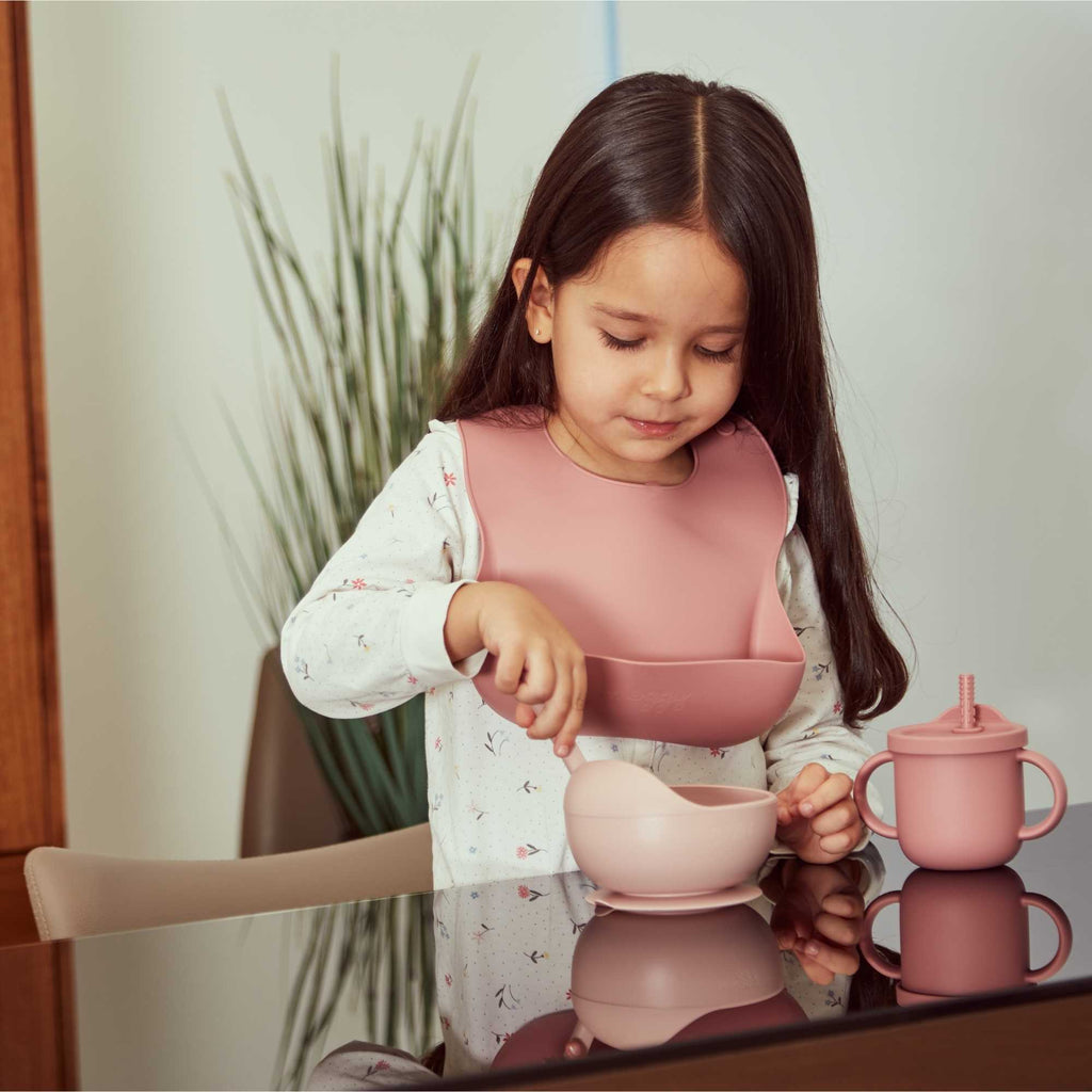 Toddler usando babero, cuchara, bowl y vaso entrenador de silicona grado alimenticio especial para alimentación complementaria color rosa marca Moggy